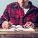 A young man wearing a checkered jacket is using a smartphone and is taking notes in a notebook at a desk
