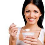 Beautiful young smiling woman holding a spoon with sour cream while standing against white background