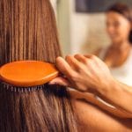 Back view of beautiful young woman in white undershirt combing her hair and smiling while looking into the mirror in bathroom