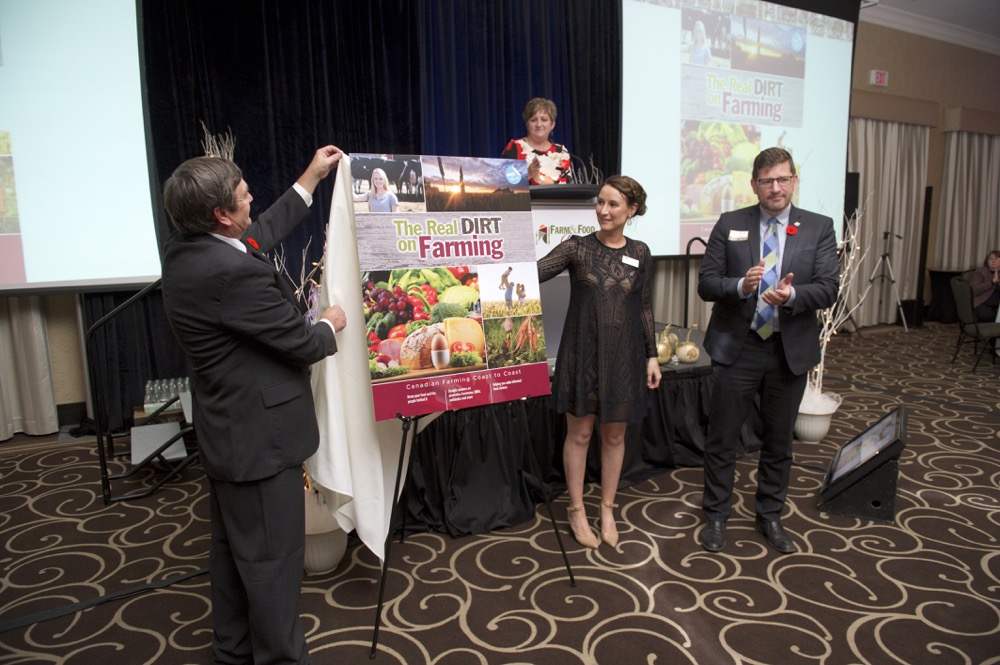 The new cover of the Real Dirt on Farming was unveiled at the Farm & Food Care Harvest Gala last week. Doing the unveiling was Farm & Food Care chair Brian Gilroy, left, Christine Schoonderwoerd, vice chair, and representing funder Wallenstein Feed & Supply Ltd., Pierre Petelle, president of CropLife Canada. Farm & Food Care Executive Director Kelly Daynard is at the podium.  Photo courtesy Farm and Food Care.
