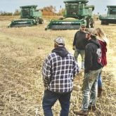 Group of people standing outside on farm field with agricultural machinery in background during harvest season