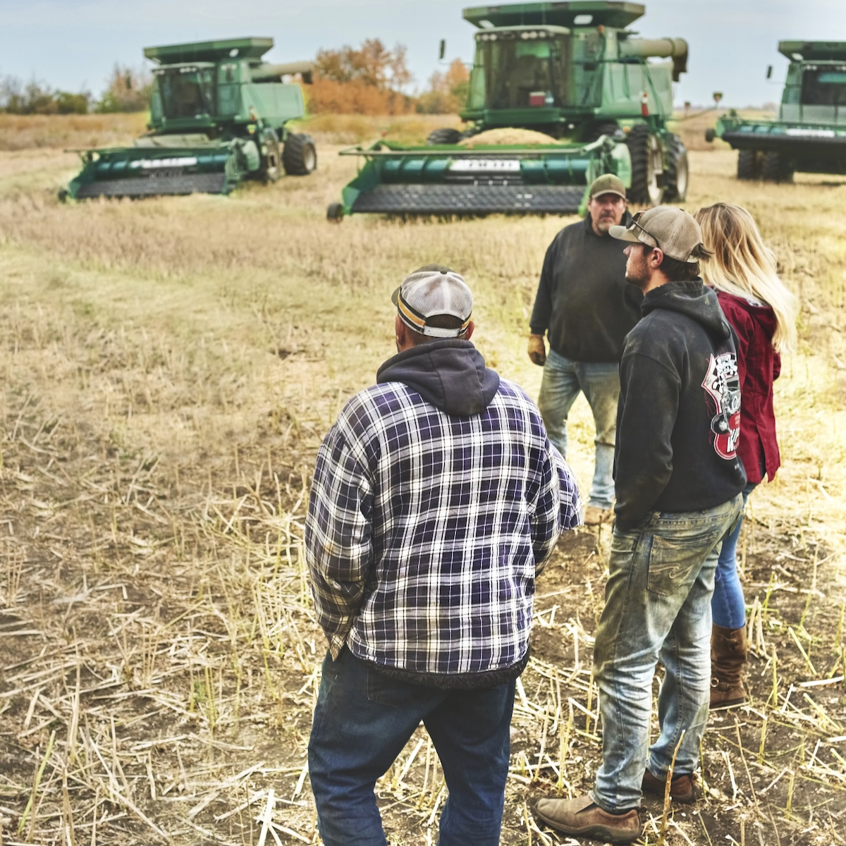 Group of people standing outside on farm field with agricultural machinery in background during harvest season