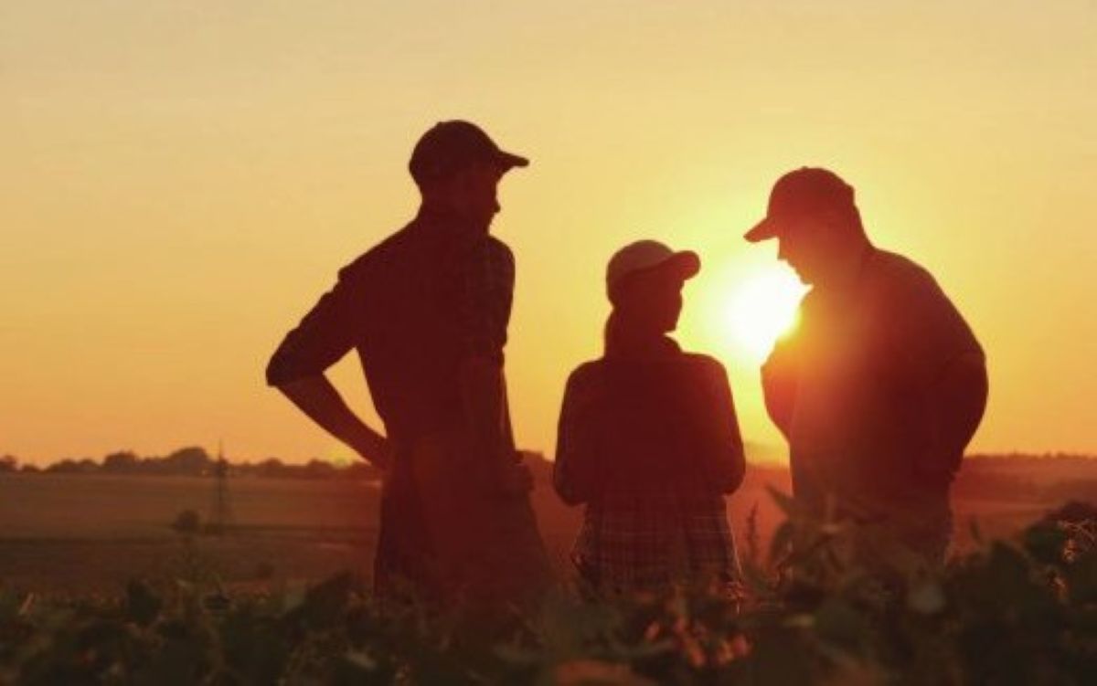 three farmers standing in a field talking silhouetted against a sunset in the background