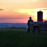 Farmer gazing out at his crop with grain silo and sunset in background.