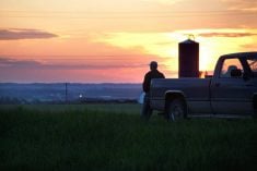 Farmer gazing out at his crop with grain silo and sunset in background.