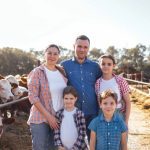 A young happy family of farmers in a cowshed. A smiling woman, a man and three kids in casual clothes take care of the pets on the farm. Mom and dad with their children in the barn